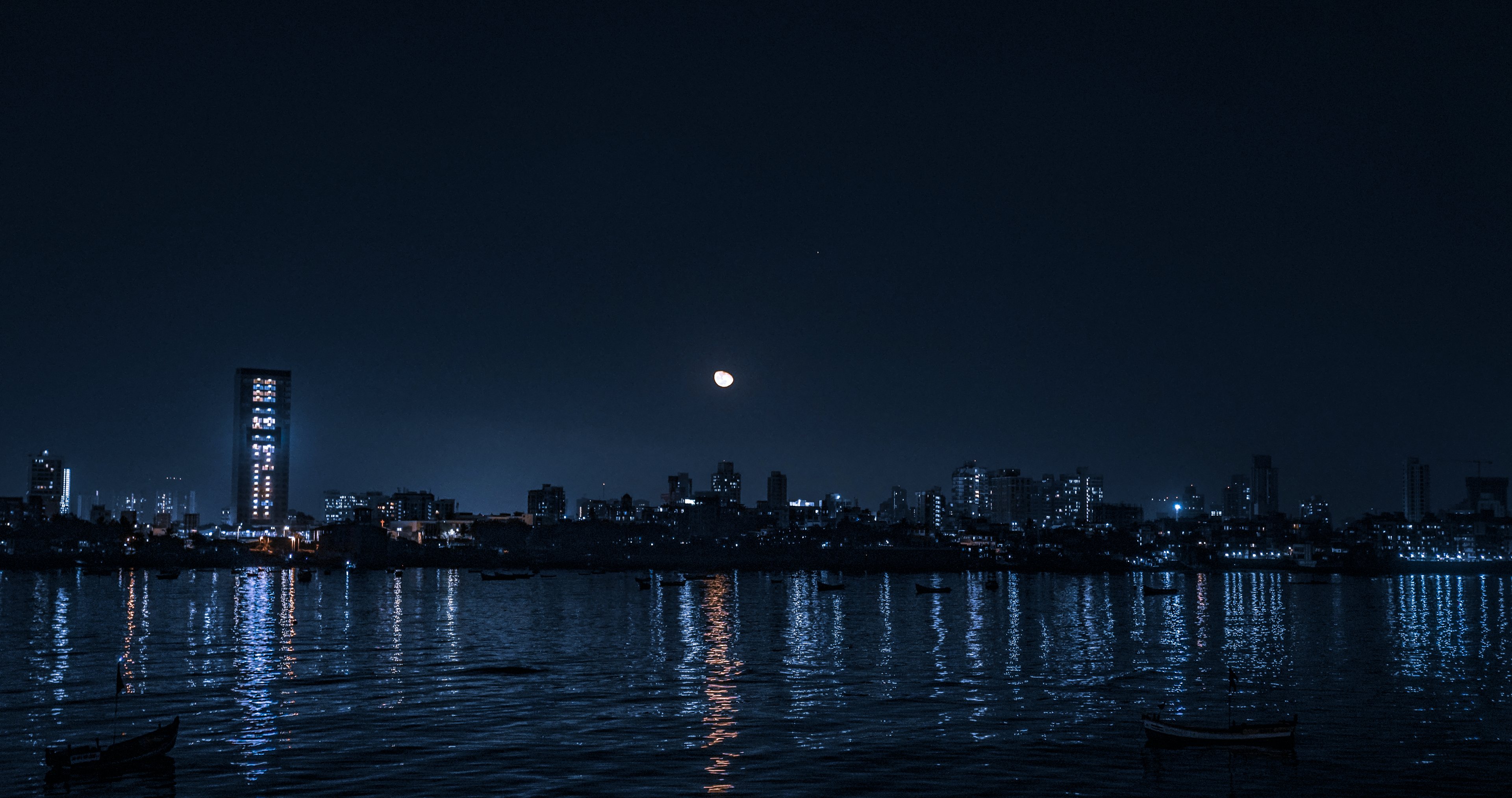 The Mumbai skyline at night across still water, a lit tower and the moon reflected in the bay